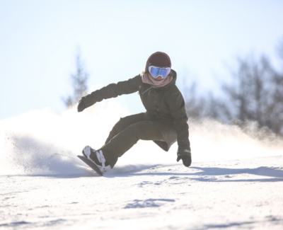A snowboarder carving through fresh snow on a bright winter day, captured mid-movement against a clear sky.